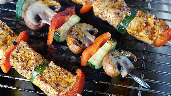 Das Bild zeigt drei Grillspieße. Der erste und dritte der Grillspieße sind abwechselnd mit Gemüse (Paprika und Zucchini) und Fleisch bestückt. Auf dem mittleren Spieß sind abwechselnd Zucchini, Pilzen und Paprika aufgespießt. Es handelt sich auf Holzspieße, die auf einem Grillrost liegen und gut gewürzt bereits leicht angebräunt sind. 